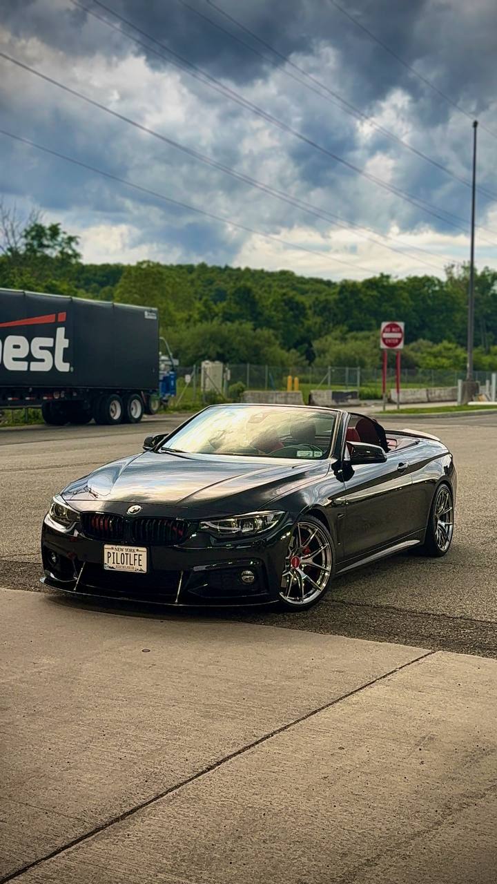 Front three-quarter of the founder's black BMW 440i convertible under a storm sky — red calipers behind polished multi-spoke wheels