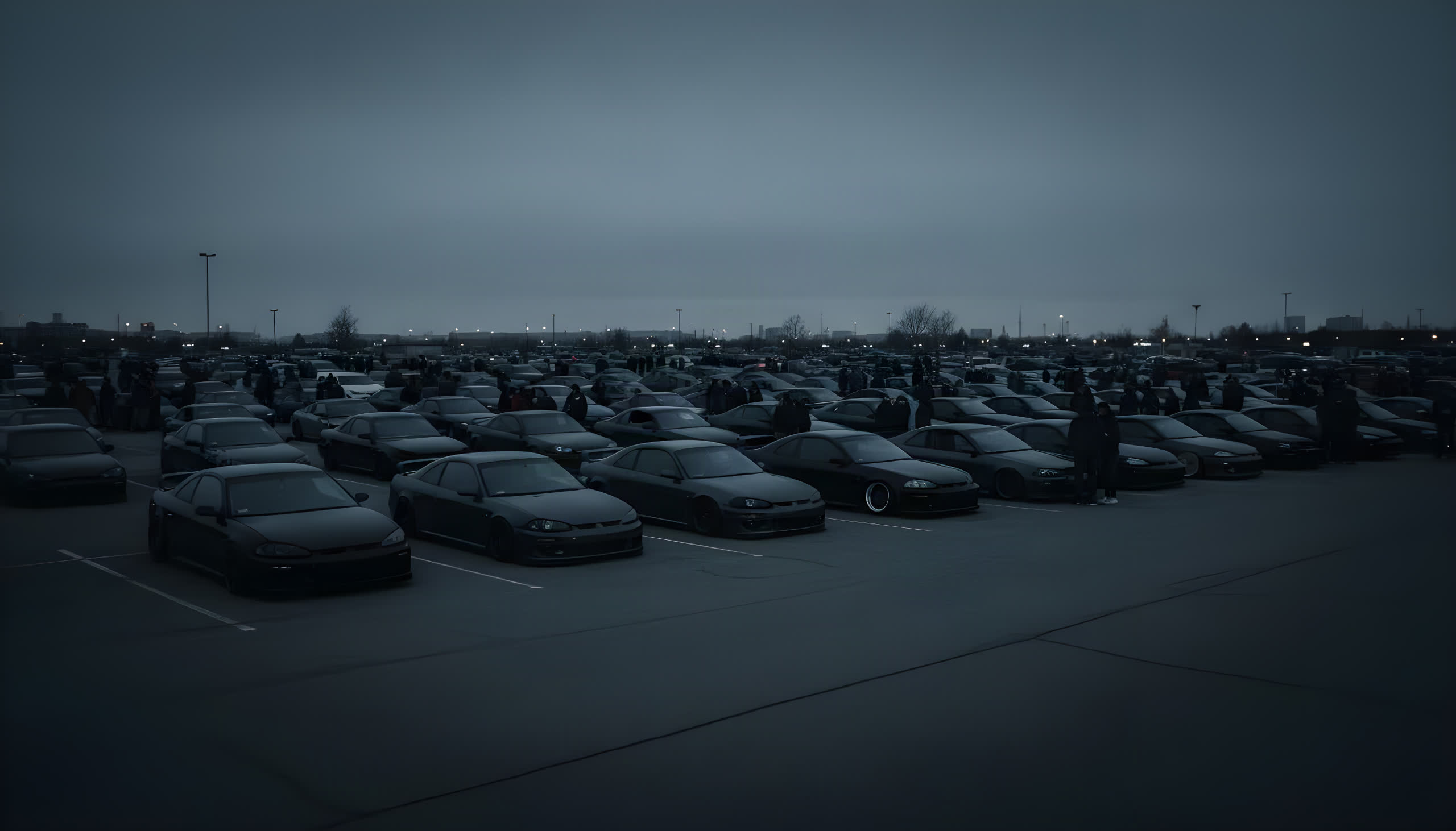 A packed night car meet — rows of low dark cars filling a lot under an overcast sky